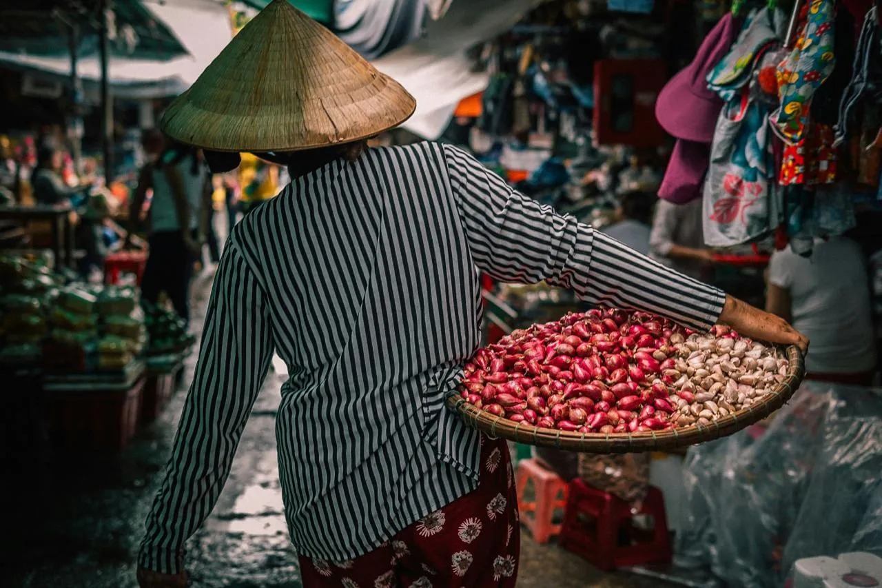 Donna con cesta di verdura in un mercato del Vietnam Donna con cappello conico che porta una cesta di verdura in un mercato vietnamita