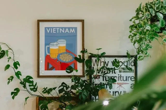 Un poster illustrato appeso a una parete tra piante verdi raffigura due boccali di bia hoi e un piatto di arachidi con la scritta Vietnam