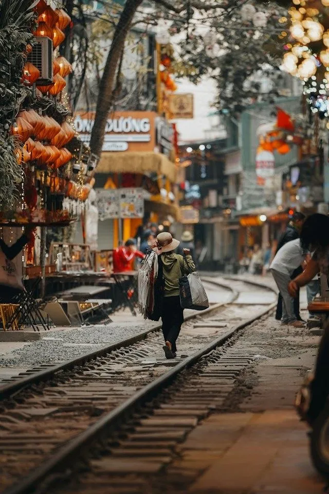 Donna con cappello conico che cammina sui binari della Hanoi Train Street, tra lanterne e caffè del quartiere vecchio