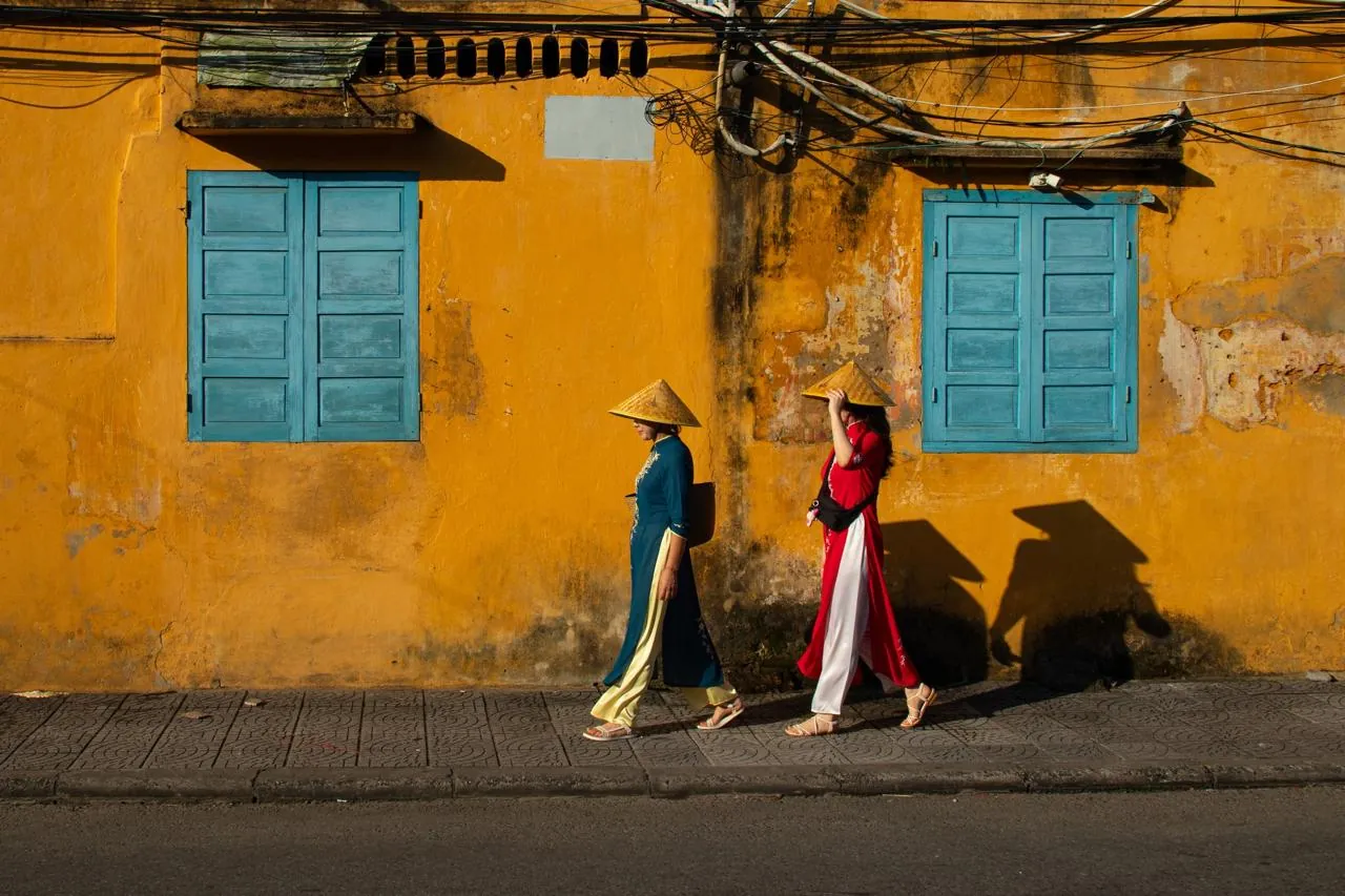 Donne in ao dai per le strade di Hoi An Due donne vietnamite in ao dai che camminano per una strada di Hoi An con muro giallo e persiane azzurre